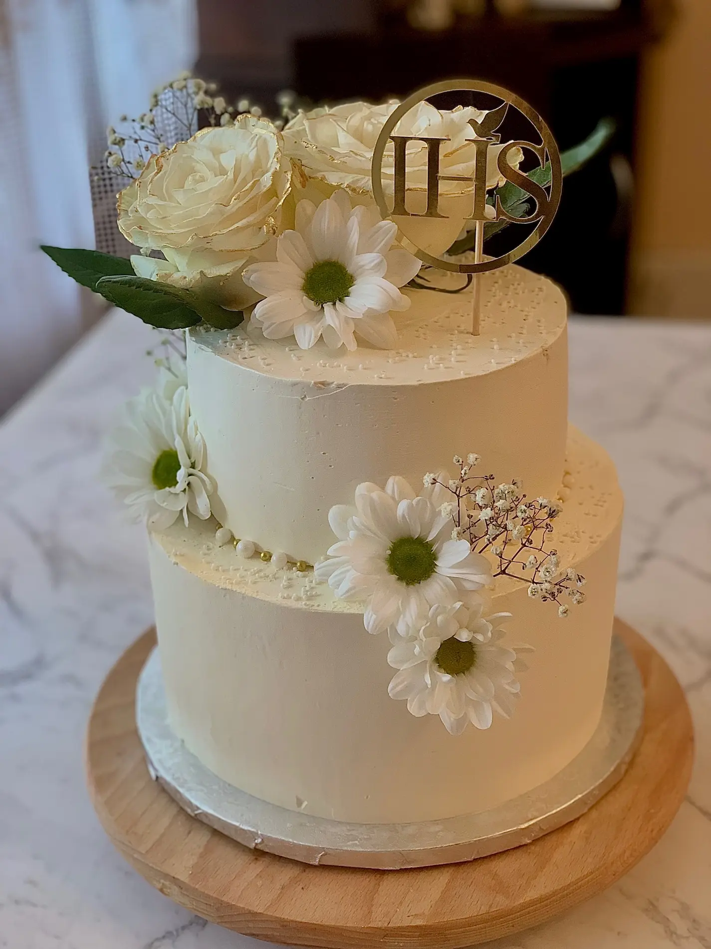 Dwupiętrowy tort komunijny ze złotym symbolem IHS, białymi stokrotkami i delikatnym kremem